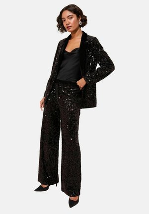 Woman wearing a black sequined blazer and matching wide-leg pants, black satin cami, black pointed heels, and silver hoop earrings.