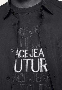 Schwarzes Hemd mit Knöpfen über einem schwarzen grafischen T-Shirt mit der Aufschrift „ACE JEANS FUTURE“ in verschiedenen Schriftarten und Texturen, mit einem glatten Stoffaussehen.