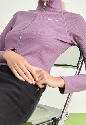 Persoon draagt een lavendelkleurige Nike-top met rits en zwarte broek, zittend op een metalen stoel met handen op schoot.