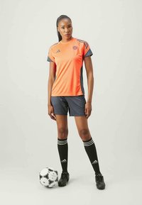 Αθλητική ενδυμασία Adidas που περιλαμβάνει μια πορτοκαλί και ναυτική μπλούζα, ναυτικά σορτς, μαύρες κάλτσες ως το γόνατο με λευκές ρίγες και μαύρα ποδοσφαιρικά παπούτσια.