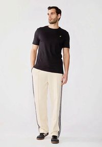 Mand står med hænderne i lommerne iført sort t-shirt, beige bukser med sorte striber og sorte sneakers.