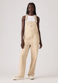 Beige denim overalls with wide legs, adjustable straps, and metal hardware. Styled with a fitted white tank top and light sneakers.