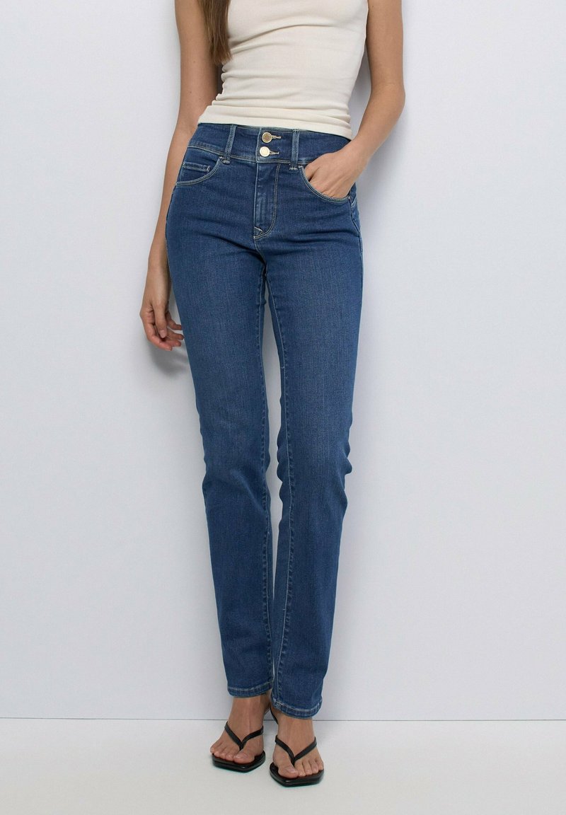 Woman wearing high-waist blue jeans with double button closure, white sleeveless top, and black square-toe flip-flop sandals standing against white wall.