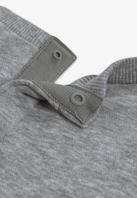 Γκρι ύφασμα με υφή φινίρισμα· διαθέτει γιακά με δύο κουμπιά κλεισίματος, με διακοσμητικές πράσινες λωρίδες κουμπιών στην κορυφή.