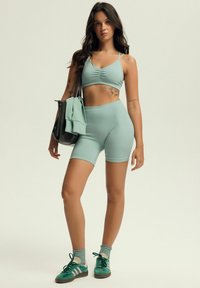 Zielono-miętowy zestaw sportowy z prążkowanego materiału, składający się z krótkiego topu z regulowanymi ramiączkami oraz szortów z wysokim stanem, w komplecie z czarną torbą i zielonymi sneakersami.