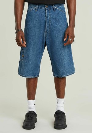 Denim-Shorts in lockerer Passform, blaue Farbe, knielang, mit vorderen Taschen und einer seitlichen Cargo-Tasche. Getragen mit schwarzen Sneakers und weißen Socken.