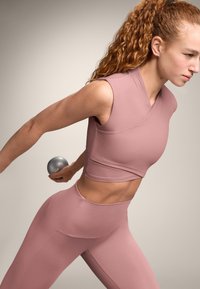 Set de echipament sportiv roz deschis, format dintr-un top fără mâneci cu textură striată și colanți cu talie înaltă. Model ținând o ganteră gri.