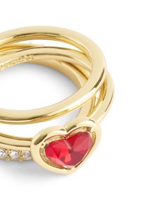 Gold-tone ring set featuring three intertwined bands. One band has a red heart-shaped stone with a faceted surface, accented by small clear stones.