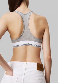 Sujetador deportivo gris con diseño de espalda nadadora y banda elástica blanca con el logo de 'Calvin Klein'. Modelo viste pantalones blancos, mostrando la vista trasera.