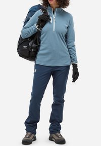 Haut à manches longues bleu clair avec demi-zippé, assorti à un pantalon bleu foncé. Les accessoires comprennent des gants noirs et un sac à dos noir avec des accents gris.