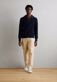 Ναυτικό πλεκτό πουλόβερ με μισό φερμουάρ, συνδυασμένο με μπεζ παντελόνι και άσπρα αθλητικά παπούτσια, σε ουδέτερο φόντο.