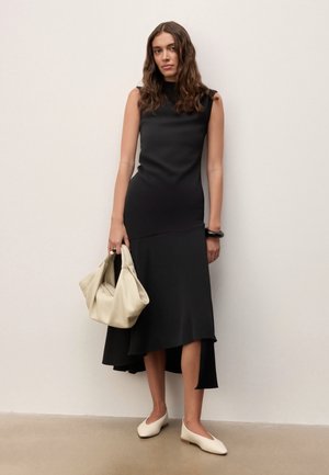 Femme portant une robe noire asymétrique sans manches, tenant un grand sac à main beige, portant des ballerines blanches et un bracelet noir, debout contre un mur blanc.