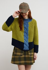 Femme souriante, portant un pull en maille  épaisse vert, marine et bleu, une jupe marron à carreaux, et une casquette marron à carreaux, tenant la visière de la casquette.