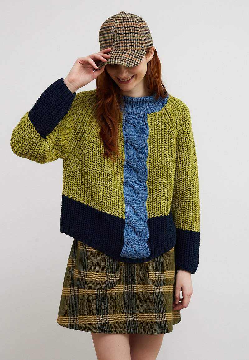 Femme souriante, portant un pull en maille épaisse vert, marine et bleu, une jupe marron à carreaux, et une casquette marron à carreaux, tenant la visière de la casquette.