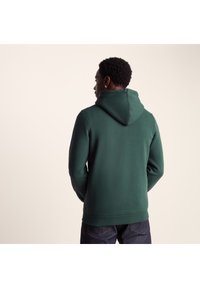 Green hoodie made from soft fabric, featuring a hood and ribbed hem. Back view shows smooth texture and structured fit.