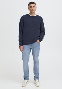 Temno modra pletena majica z rebrasto teksturo, z okroglim izrezom, kombinirana s svetlo modrimi bombažnimi kavbojkami in bež supergami. Preprost, priložnosten videz.