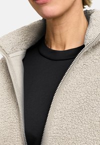 Giacca in pile con zip di colore beige chiaro, con superficie texturizzata e colletto morbido, indossata sopra un top nero a collo alto. Dettagli della zip visibili.