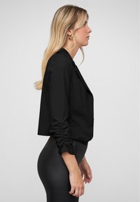 Černý zkrácený blazer se strukturovaným designem a nařasenými rukávy. Spárován s černými legínami s vysokým pasem. Jednoduchý, elegantní vzhled.