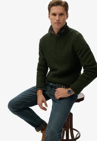 Pull en maille vert foncé avec une finition texturée, porté par-dessus une chemise à carreaux, associé à un jean bleu et des bottines marron à lacets. Montre au poignet.