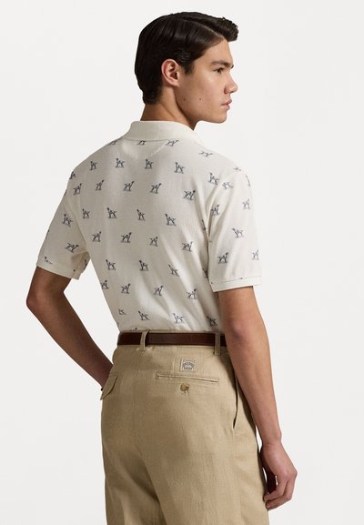 Kortekrige poloshirt in crème met een blauw hondenpatroon; gedragen met beige broeken met een achterzak en knoopdetail.