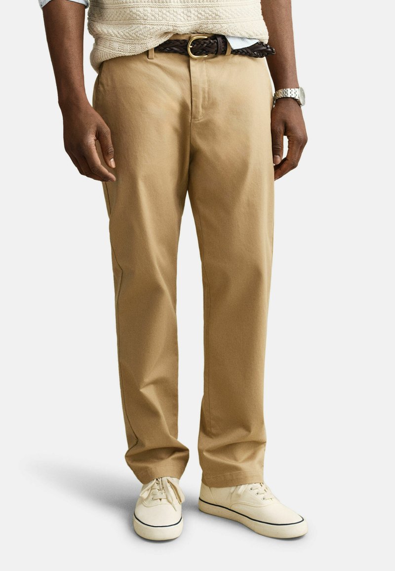 Hombre con pantalones chinos beige, zapatillas blancas, un cinturón trenzado marrón, un chaleco de punto crema y un reloj de pulsera plateado, de pie frente a un fondo blanco.