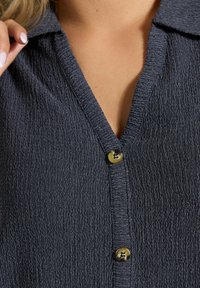 Granatowa bluzka o fakturowanej powierzchni z dekoltem w serek, krótkim kołnierzykiem i pięcioma guzikami. Materiał ma zmarszczoną fakturę i delikatny połysk. Dodatki w postaci guzików są złote.