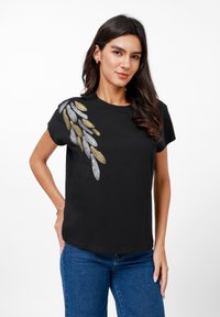 T-shirt en coton noir avec un motif de feuilles brodé en métal argenté et doré sur l'épaule gauche, manches courtes et coupe décontractée.
