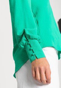 Grüne Bluse mit langen Ärmeln aus strukturiertem Stoff. Mit gerafften Ärmeln und drei passenden Knöpfen aus demselben Stoff am Bündchen. Kombiniert mit weißen Hosen.
