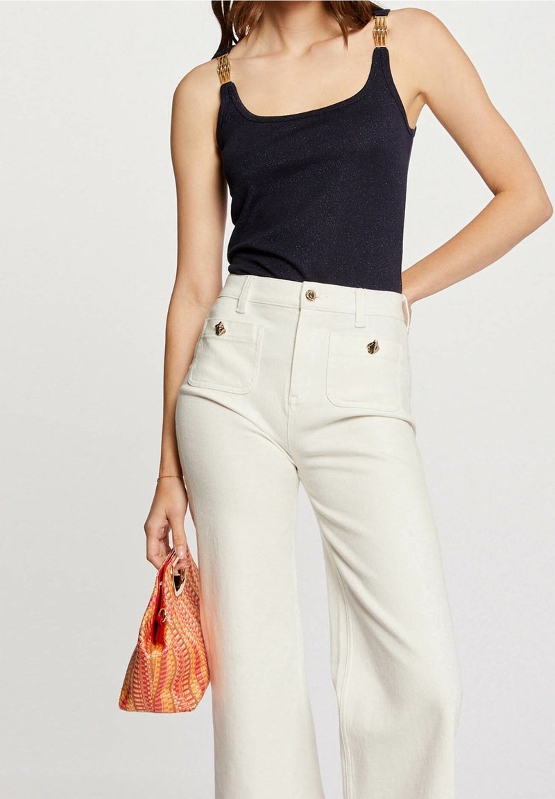 Femme portant un haut noir sans manches avec des détails dorés, un pantalon blanc taille haute, et tenant un petit sac à main orange à motifs.