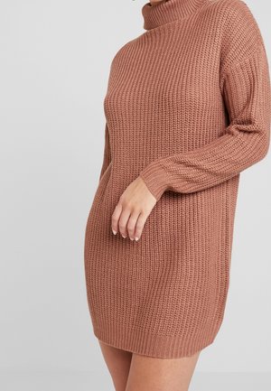 Frau trägt ein langes, grob gestricktes, staubig-rosafarbenes Rollkragen-Strickkleid, mit einer Hand, die nahe der Hüfte an einem schlichten Hintergrund ruht.