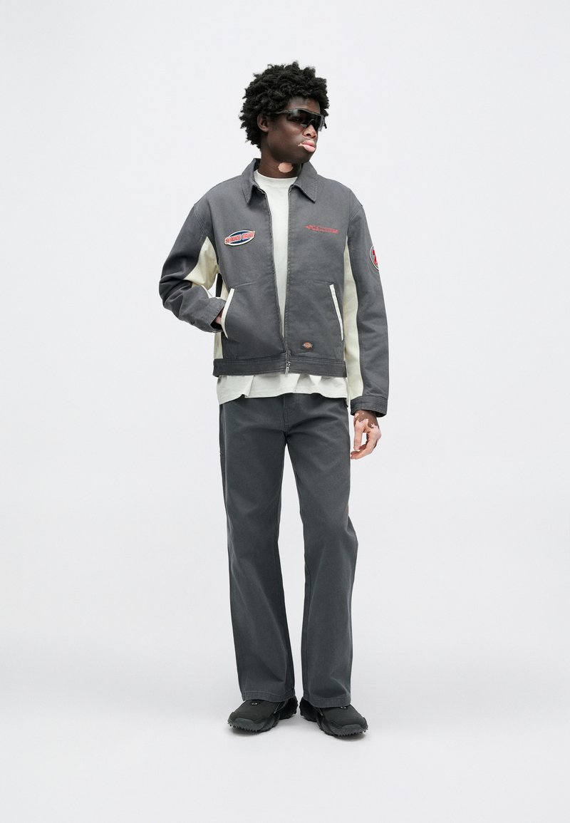 Gray and cream jacket with embroidered patches, front zipper, side pockets; paired with gray pants and dark shoes. Model wears sunglasses.