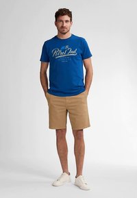 Blaues Baumwoll-T-Shirt mit "Petrol Ind"-Grafik, kombiniert mit khakifarbenen Shorts. Das Model trägt weiße Sneaker; einfarbiger Hintergrund.