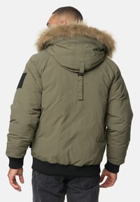 Olivgrüne Kapuzen-Puffermantel mit Kunstfellbesatz, gerippten Bündchen und einem strukturierten schwarzen Patch am Ärmel. Verfügt über einen Reißverschluss.