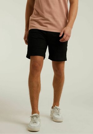 Homme portant un short noir, un haut en maille beige et des baskets blanches, debout devant un fond clair uni.