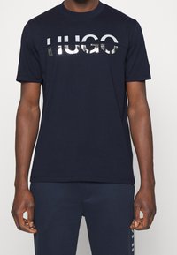 Navy blue cotton T-shirt featuring a silver reflective "HUGO" logo across the chest, crew neckline, and short sleeves.