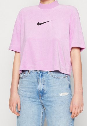 Person trägt ein kurzes lavendelfarbenes Nike-T-Shirt mit kurzen Ärmeln und eine hoch taillierte hellblaue zerrissene Jeans vor neutralem Hintergrund.