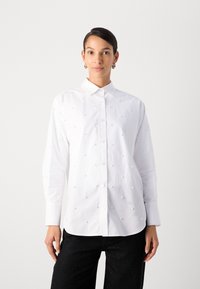 Camicia bianca con bottoni, caratterizzata da dettagli testurizzati, arricchita da piccole decorazioni a forma di perla e con colletto classico, abbinata a pantaloni neri.
