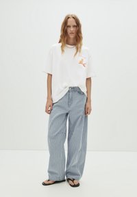 Weißes, übergroßes T-Shirt mit einem bunten Grafikdesign und lässigen, blau-weißen gestreiften weiten Hosen. Schwarze Flip-Flops an nackten Füßen.