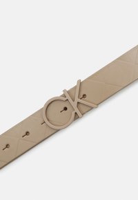 Beige leren riem met een quiltpatroon, voorzien van een ronde gesp met de letters "CK" in een bijpassende tint. Inclusief meerdere verstelgaten.