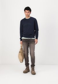 Felpa blu navy con logo in rilievo, sottoshirt verde chiaro, jeans grigi, stivaletti beige e borsa beige. Design semplice e casual.