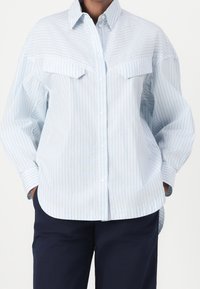 Person som bär en lös passande ljusblå och vit vertikalt randig skjorta med bröstfickor och mörkblå byxor, h änderna i fickorna.