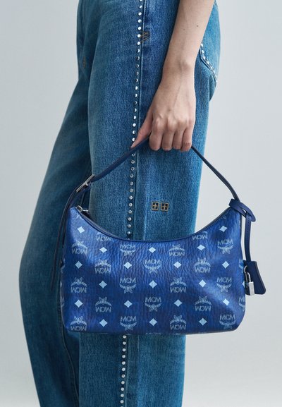 Ręka trzyma niebieską skórzaną torbę MCM z wzorem logo, obok niebieskich dżinsów z ćwiekami.