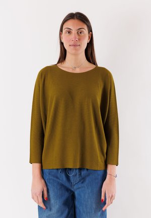 Donna con capelli castani lunghi che indossa un maglione largo verde oliva e jeans blu, in piedi davanti a uno sfondo bianco uniforme.