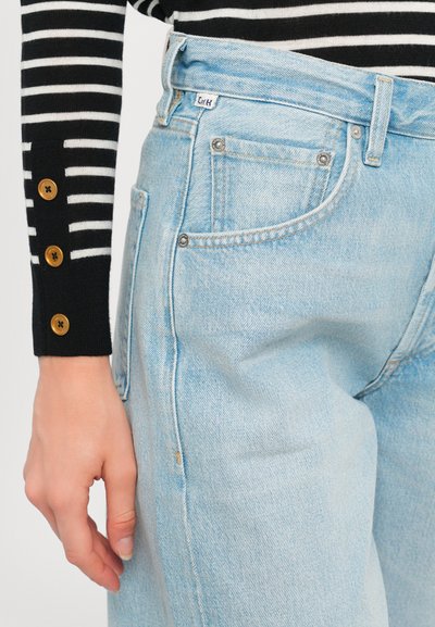 Jeans in denim azzurro chiaro con una vestibilità tapered, cinque tasche e un leggero effetto scolorito. Un maglione a righe bianche e nere presenta dettagli con bottoni sulle maniche.