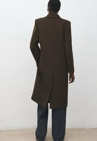 Brązowy wełniany płaszcz o dopasowanym kroju, z długimi rękawami i poduszkami na ramionach. Posiada tylny rozporek i kołnierz. W połączeniu z szerokimi niebieskimi dżinsami.