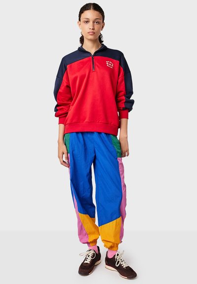 Red and navy half-zip sweatshirt with logo, paired with multi-colored (blue, green, pink, yellow) track pants and dark footwear.