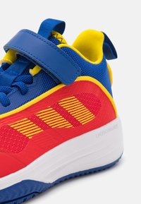 Baskets de sport rouges et bleues avec un accent jaune, featuring une tige texturée, une sangle Velcro et un motif rayé sur le côté.