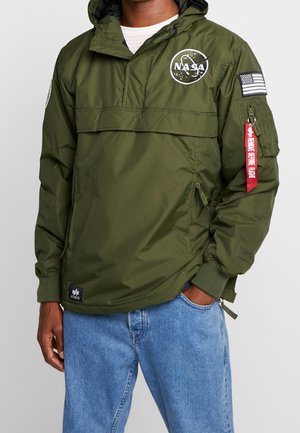 Person trägt eine olivgrüne NASA-Kapuzensjacke mit Aufnähern auf Brust und Ärmel, kombiniert mit hellblauen Jeans.