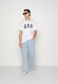 White cotton t-shirt featuring bold navy "GAP" logo, paired with light blue denim jeans and white sneakers. Accessories include black sunglasses.