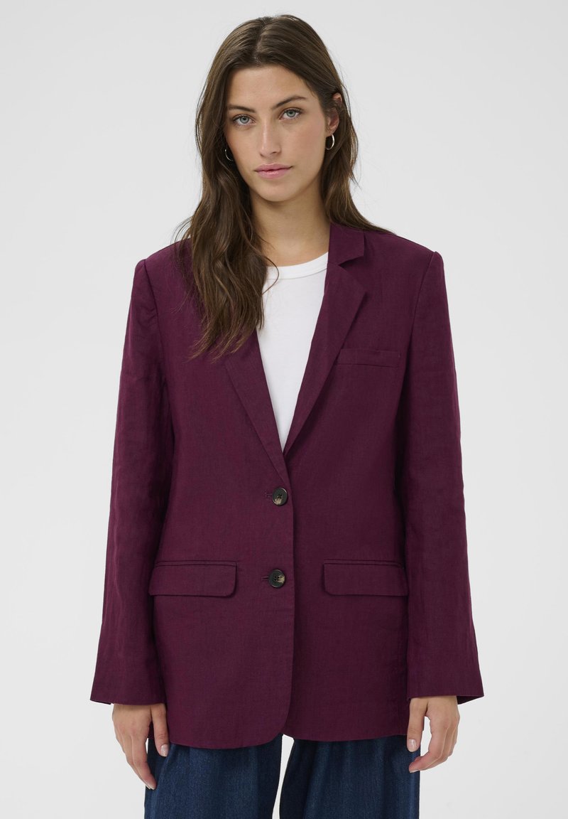 Femme aux longs cheveux bruns portant un blazer violet boutonné sur une chemise blanche et un pantalon bleu foncé, debout devant un fond uni.